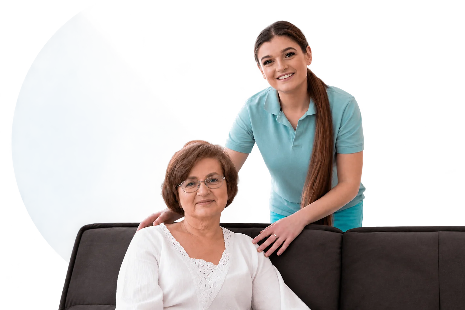 Caregiver standing behind an elderly woman on a sofa, offering support at home in a living room setting.